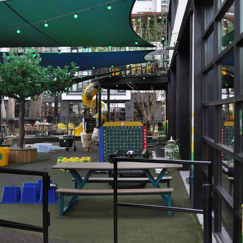 Outdoor play area with picnic tables, green shade sails, playground equipment, and large connect four game board