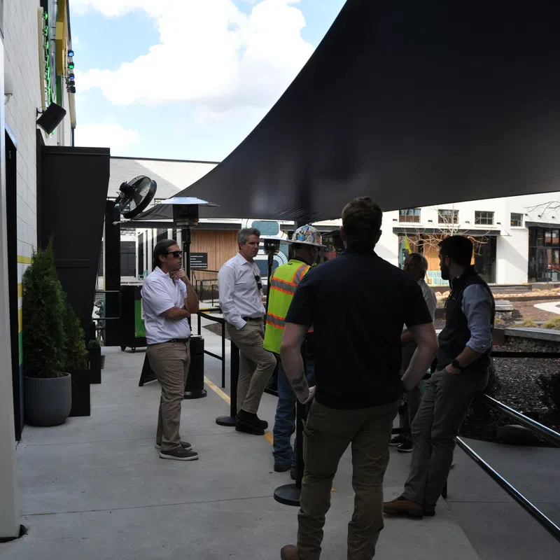 Group of people, including one in a safety vest, talking under a large black canopy outside modern buildings on a sunny day