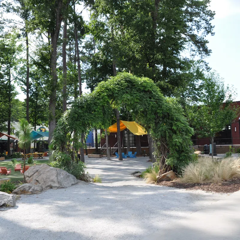 Green leafy archway at the entrance of a playground with trees, benches, and a brick building in the background.