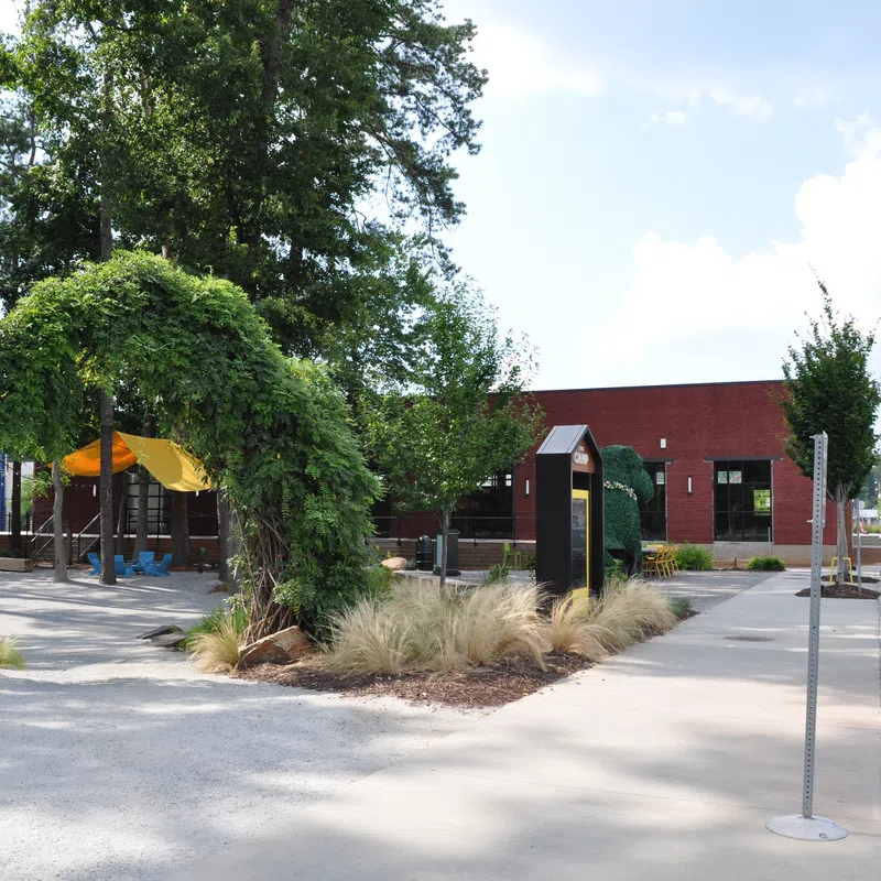 Outdoor park area with green leafy arch, playground with yellow canopy, sidewalk, and parked vehicles on a sunny day