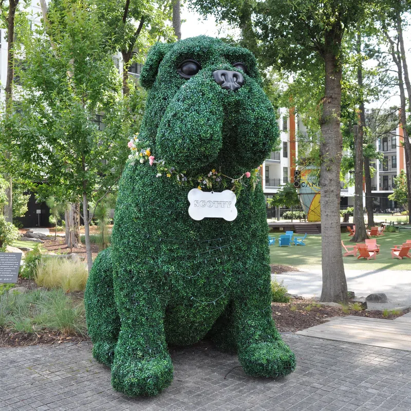 Large topiary sculpture of a dog wearing a floral collar and bone-shaped tag in an urban park setting.