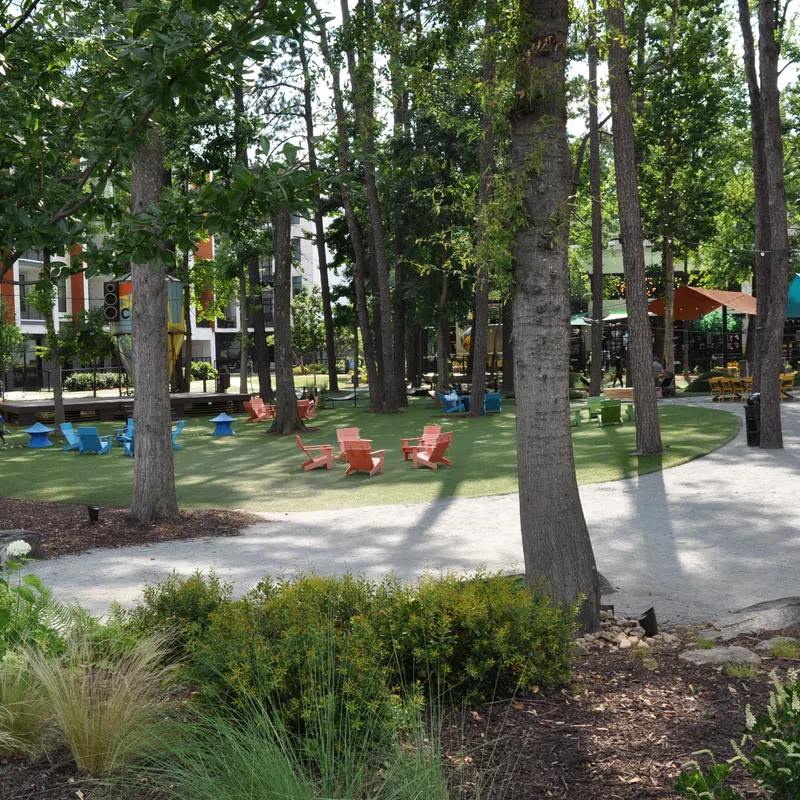 Outdoor urban park with trees, colorful chairs on green lawn, winding pathways, and shaded seating areas under canopies.