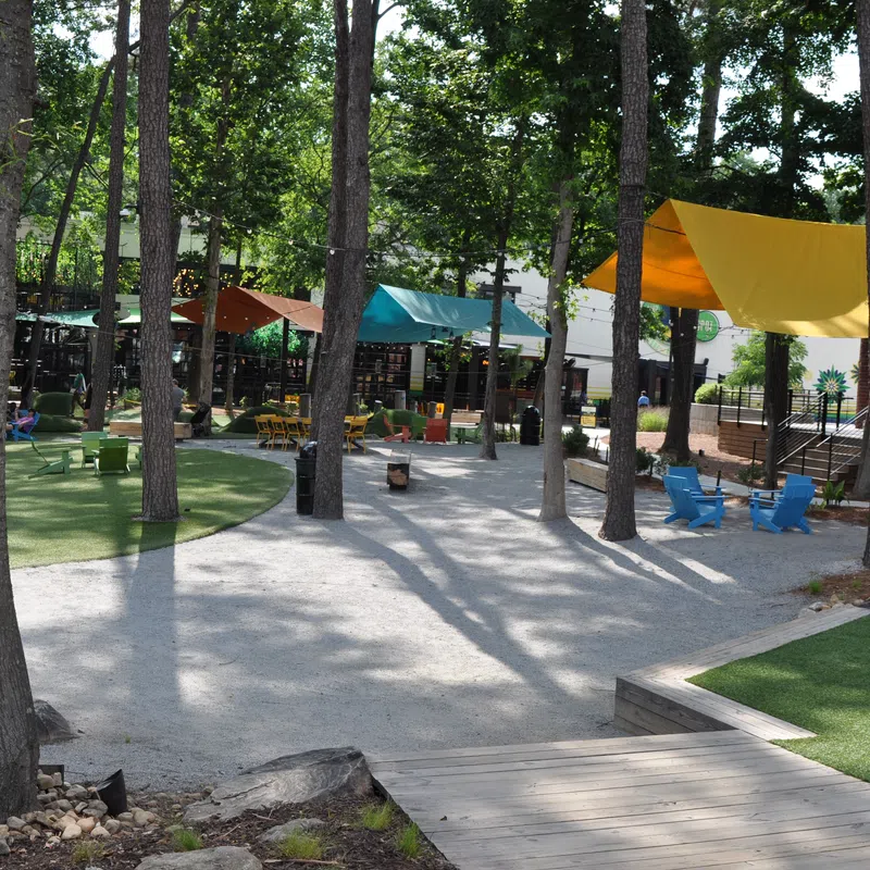 Shaded outdoor seating area with colorful chairs and canopies among trees on gravel and grass paths.