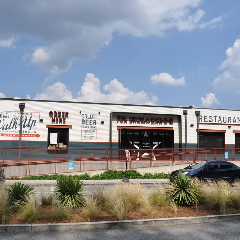 Exterior of Fox Bros Bar-B-Q restaurant with a walk-up window, outdoor seating, and parked cars on a sunny day.