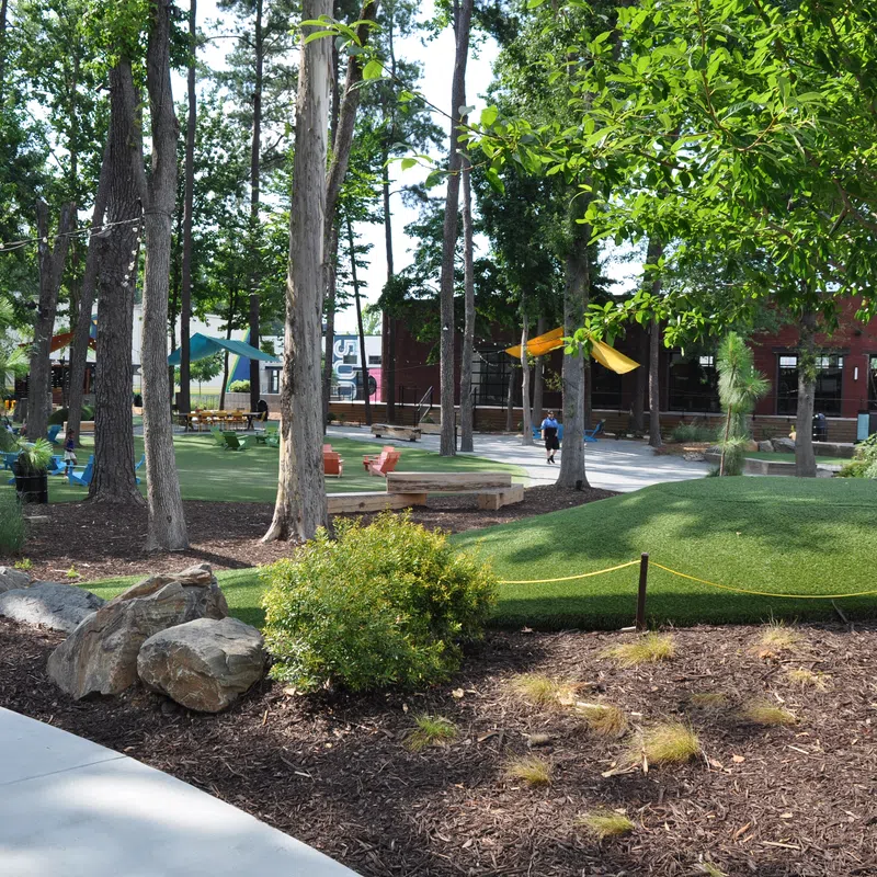 Community park with large trees, green turf, walking paths, playground equipment, and seating areas under sunny sky.