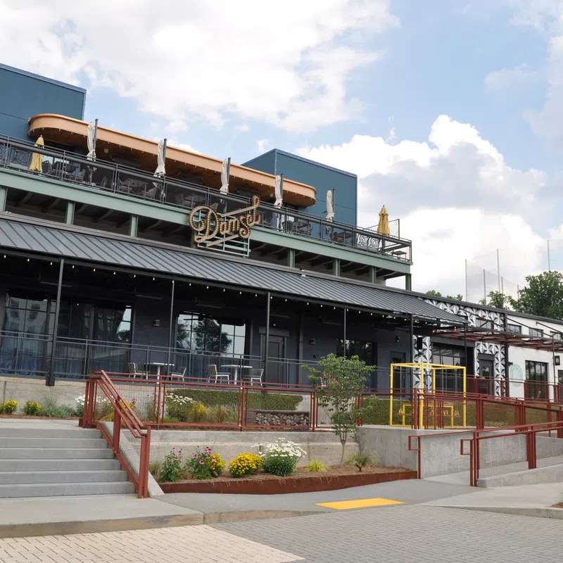 Modern restaurant building with outdoor seating, stairs, ramps, and flower beds under partly cloudy sky.