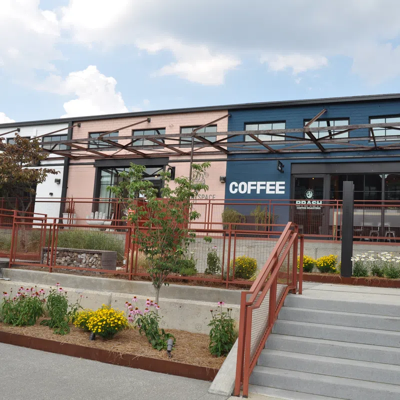 Outdoor view of a coffee shop with seating, plants, stairs, and a colorful directory sign on a sunny day.