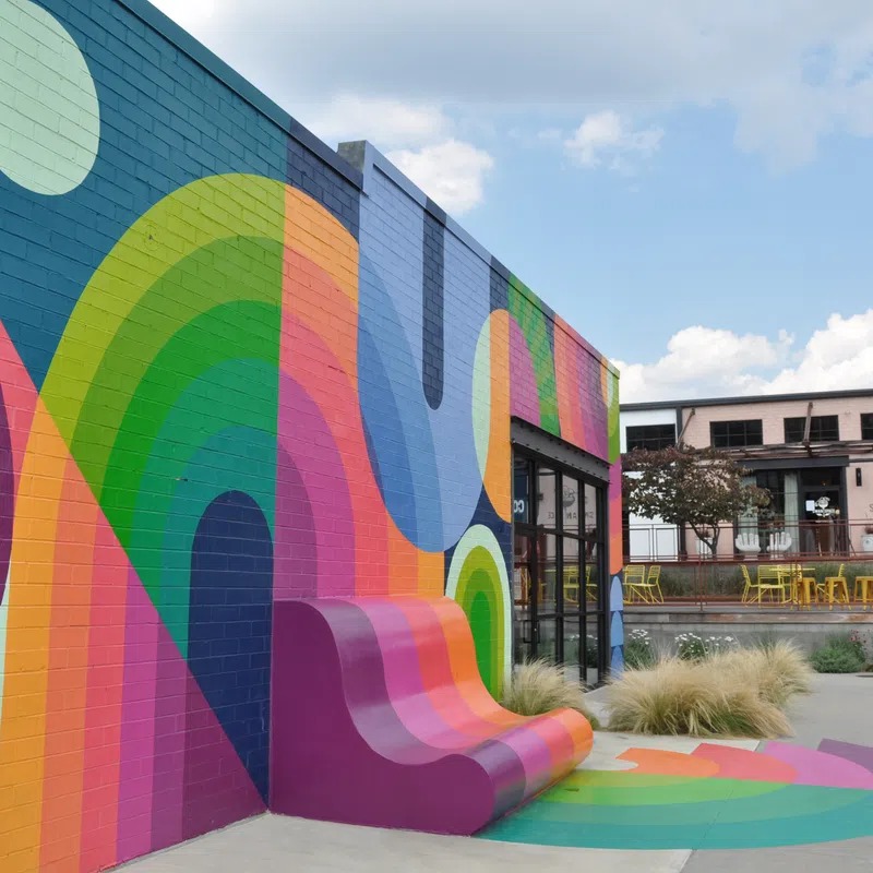 Colorful mural with vibrant rainbow patterns and curved bench on an urban building exterior under blue sky.