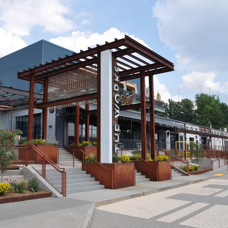 Modern building entrance with rust-colored steel pergola, concrete stairs, and landscaped planters under a partly cloudy sky