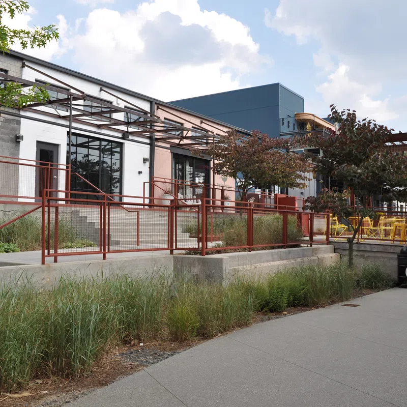 Modern building exterior with red railings, outdoor seating, trees, and a clear sky with clouds.