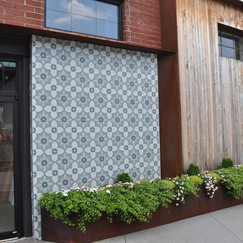 Exterior building wall with patterned tile feature, planter box with greenery and flowers, black door, and wooden siding.