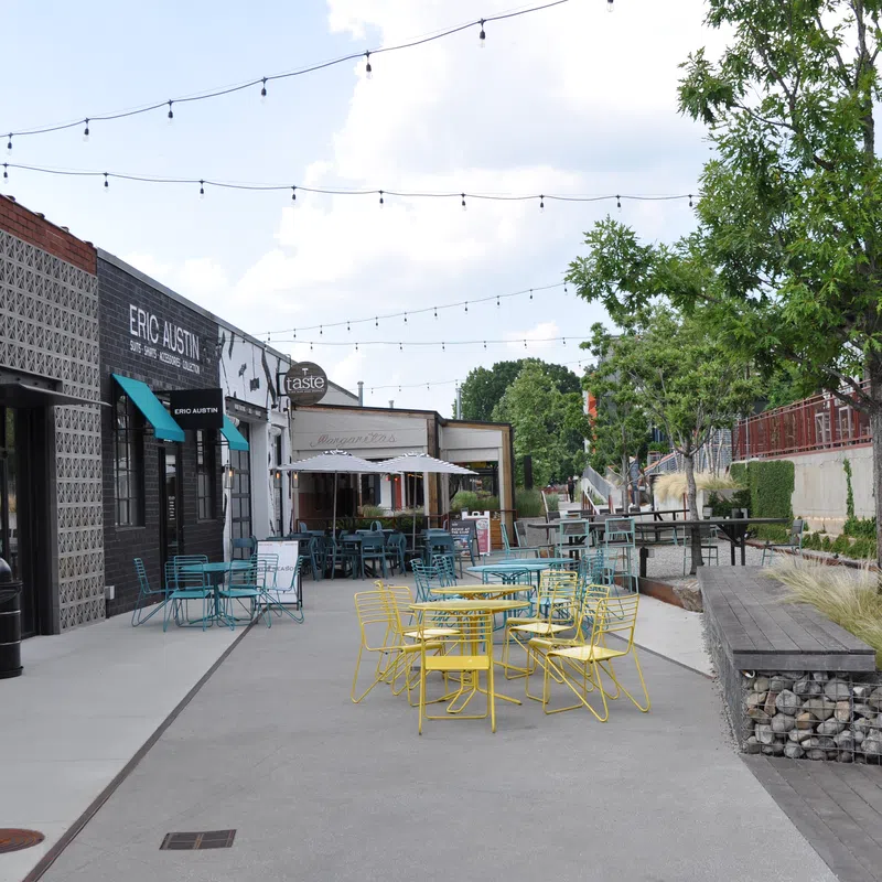 Outdoor seating with colorful chairs and tables along a sidewalk at a cafe under string lights on a sunny day.