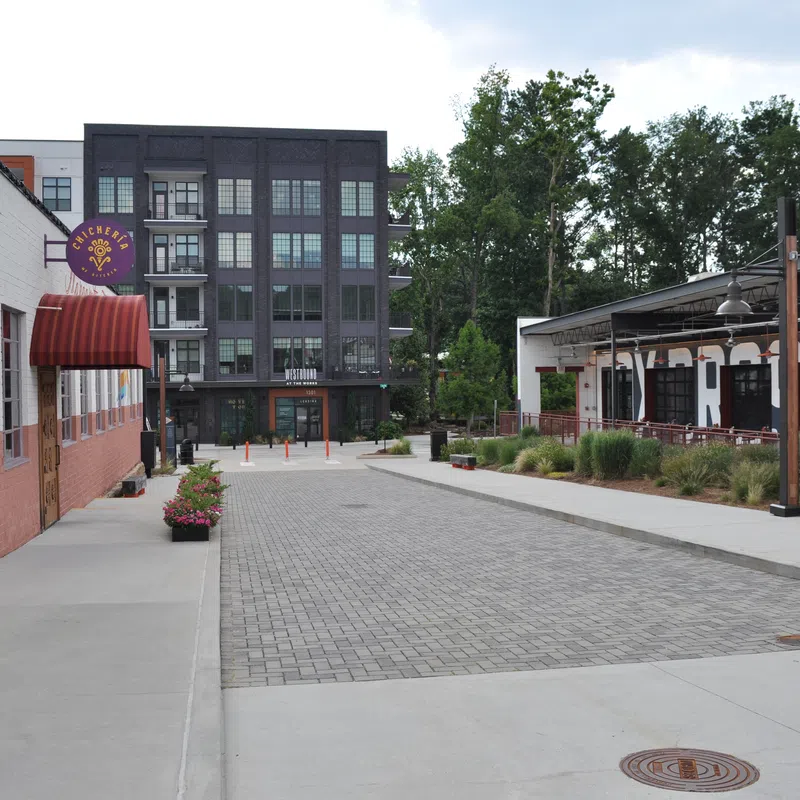 Empty urban street with modern buildings, outdoor seating, greenery, and a cloudy sky background.