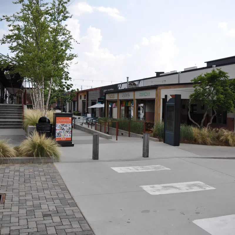 Outdoor shopping and dining area with benches, greenery, and storefronts under a partly cloudy sky in daytime.
