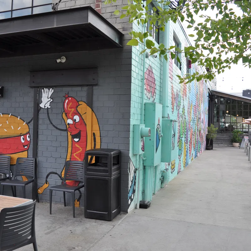 Outdoor seating area with mural of cartoon burger and hot dog high-fiving on brick wall at restaurant.