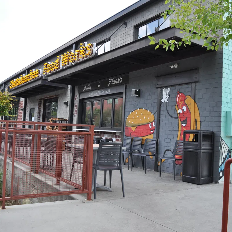 Exterior of a restaurant with murals of a cheeseburger and hot dog waving, outdoor seating, and a red metal fence.