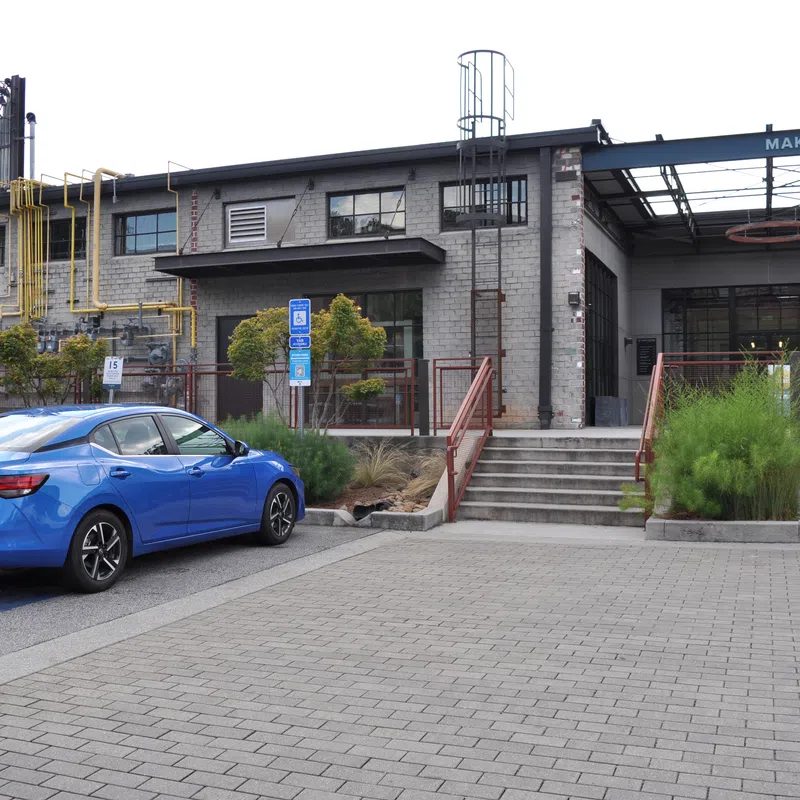 Blue sedan parked in front of a gray industrial Makers Building with stairs and greenery on a cloudy day