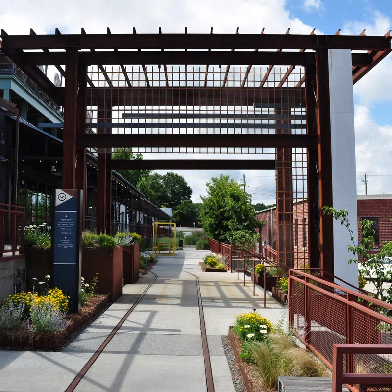 Modern outdoor walkway with metal pergola, planters, and urban buildings under a blue sky with clouds.