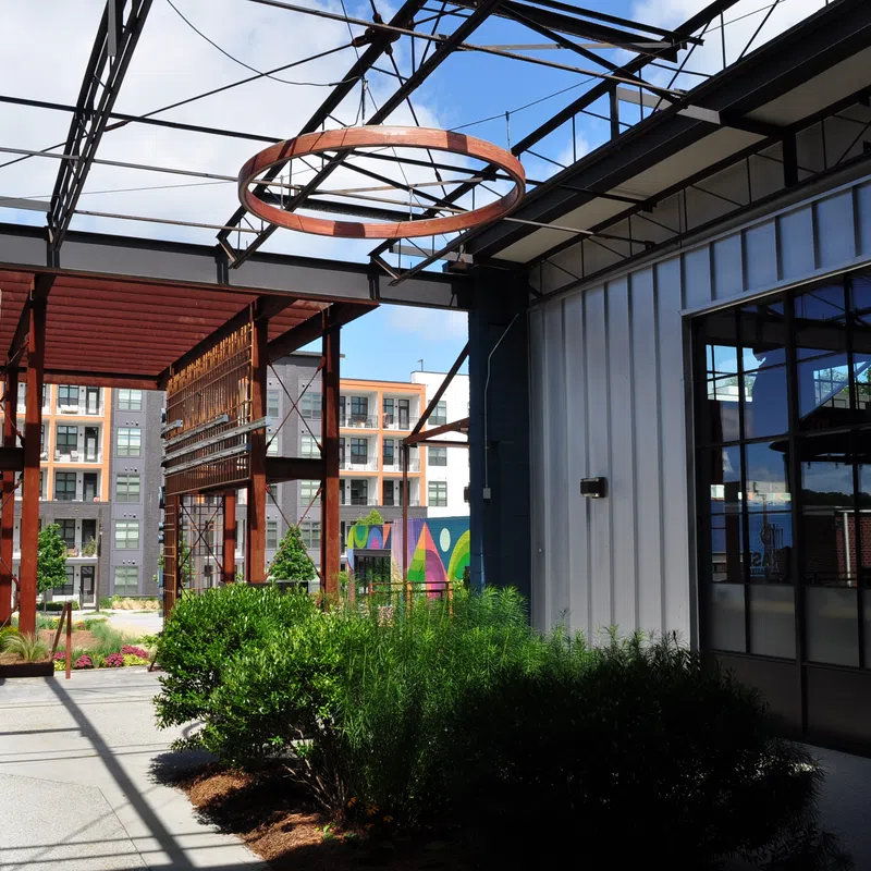 Modern industrial-style outdoor corridor with greenery, metal framework, glass windows, and apartment buildings in background