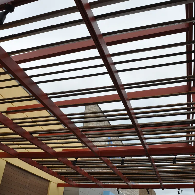 Modern pergola with wooden slats and red beams over a building exterior under cloudy sky.