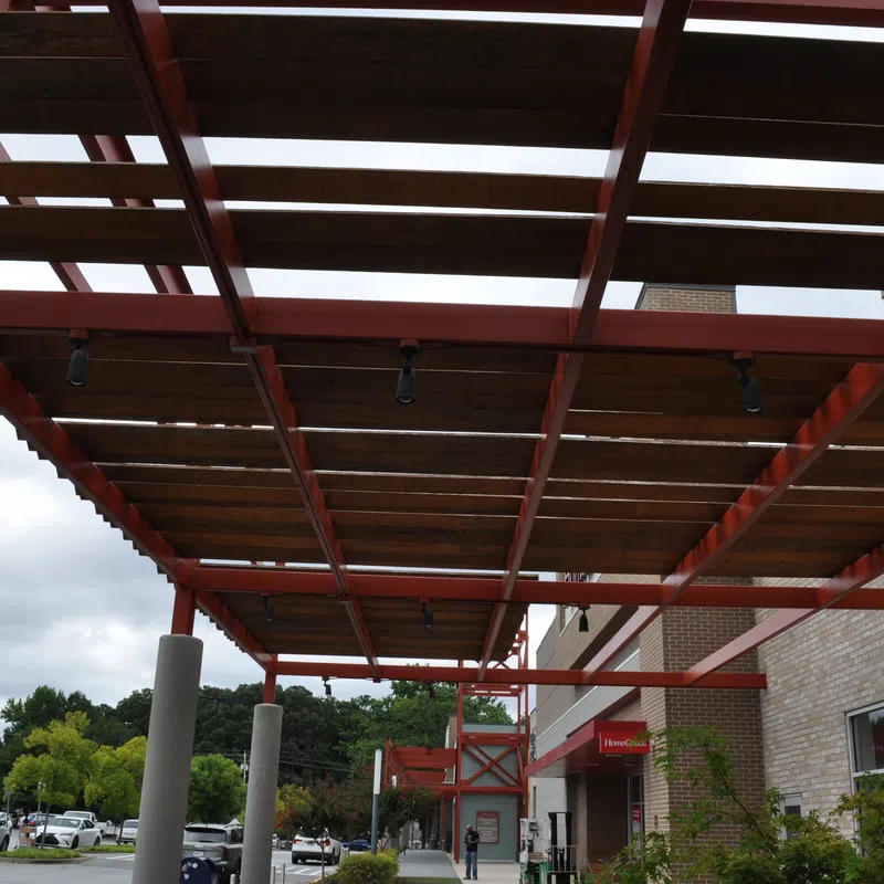 Outdoor walkway with red steel pergola and wooden slats next to a brick building under cloudy sky.