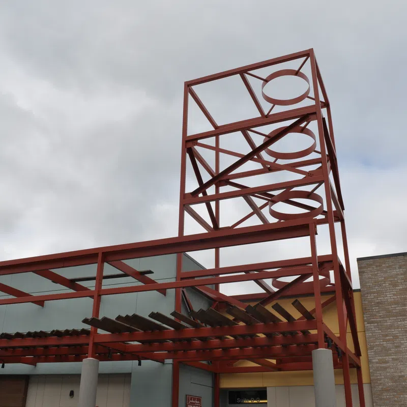 Red metal framework structure with circular cutouts above a building entrance under cloudy sky.