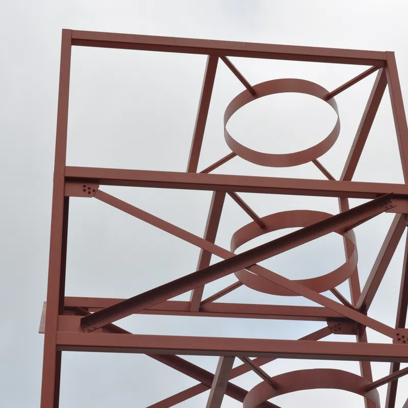 Rust-colored metal framework with circular and diagonal supports against a cloudy sky.