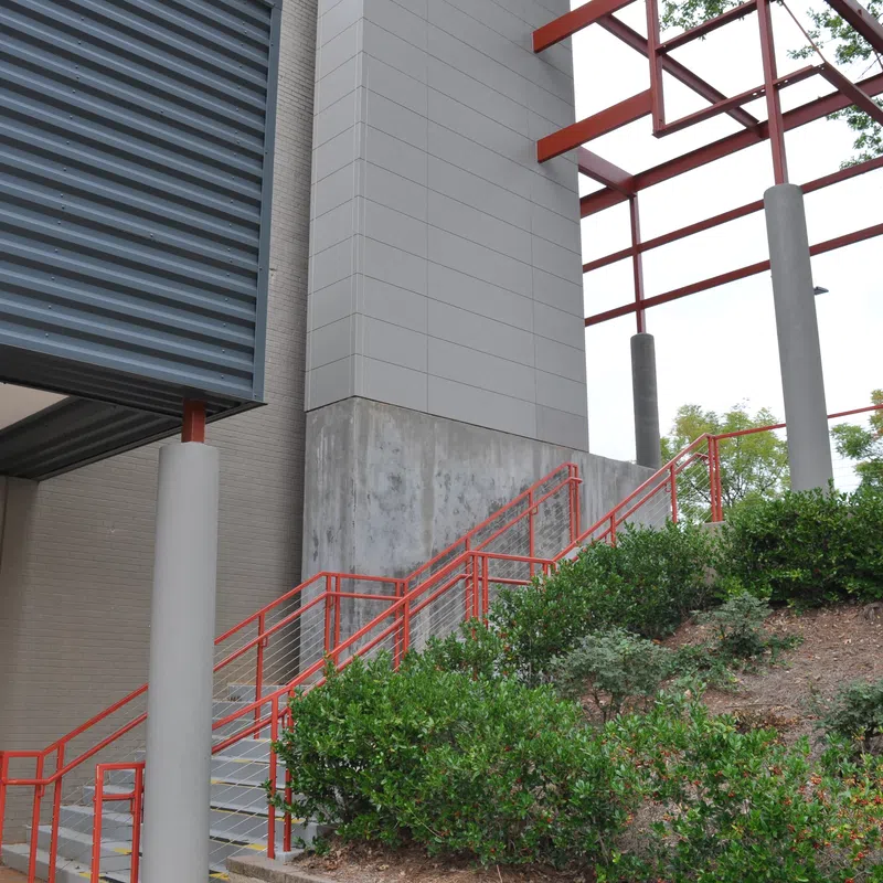 Outdoor staircase with red railings next to modern building and green shrubbery under metal canopy.