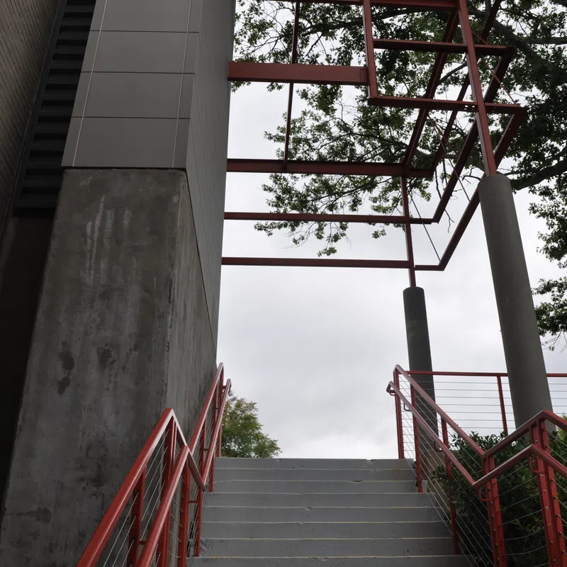 Concrete stairs with red railings lead up to a metal frame structure surrounded by trees against a cloudy sky.