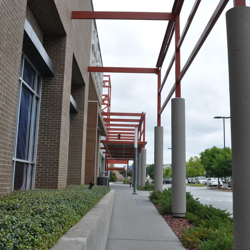 Sidewalk beside a brick building with red steel beams and concrete pillars under an overcast sky near a parking lot.