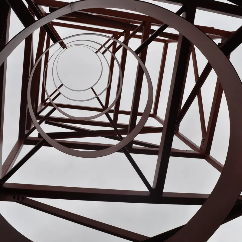 Looking upward inside a red metal tower structure with circular and diagonal beams against a cloudy sky.