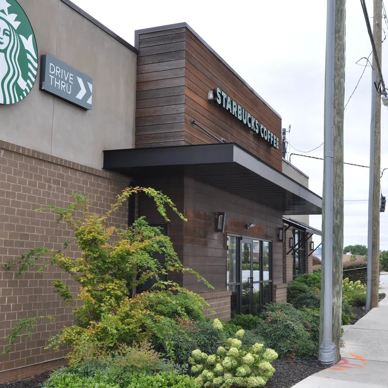 Starbucks coffee shop exterior with drive-thru sign and landscaped greenery by busy street corner.