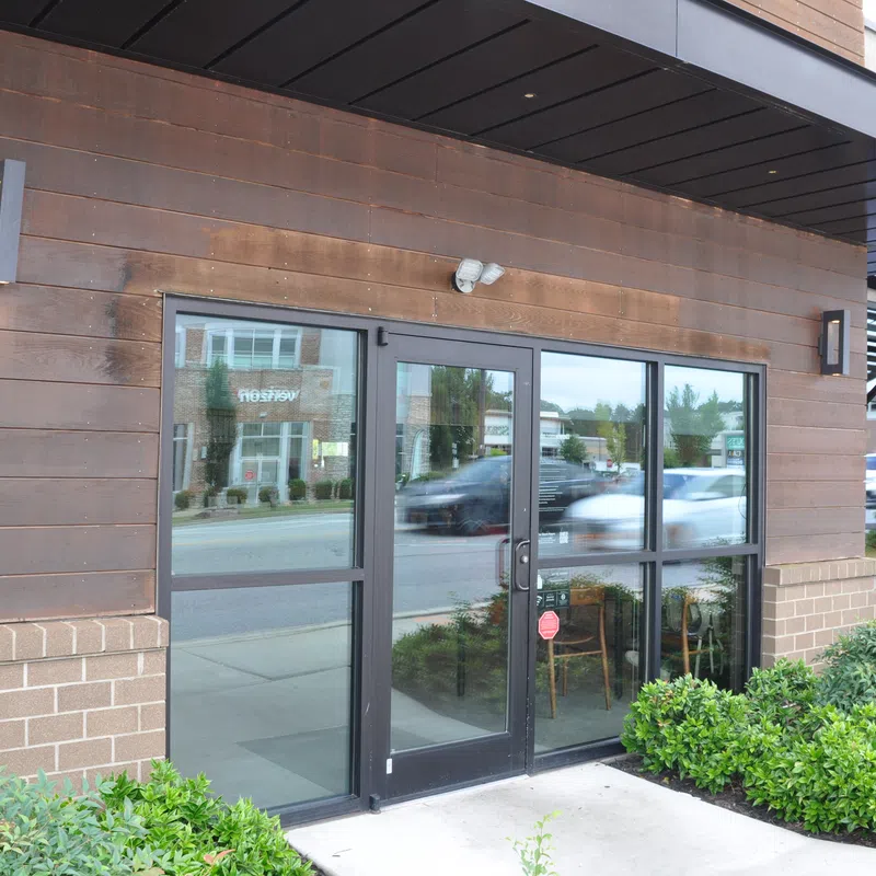 Glass double doors with black frames at storefront reflecting street and cars, surrounded by wood and brick exterior with greenery.