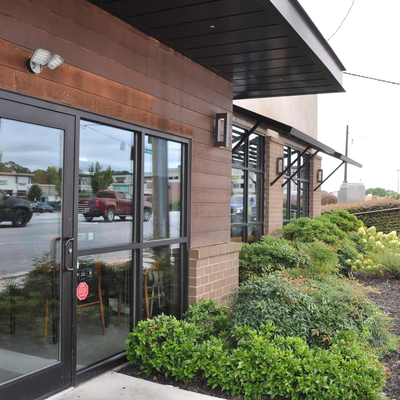 Commercial building entrance with glass doors and landscaped greenery along the sidewalk on a busy street.