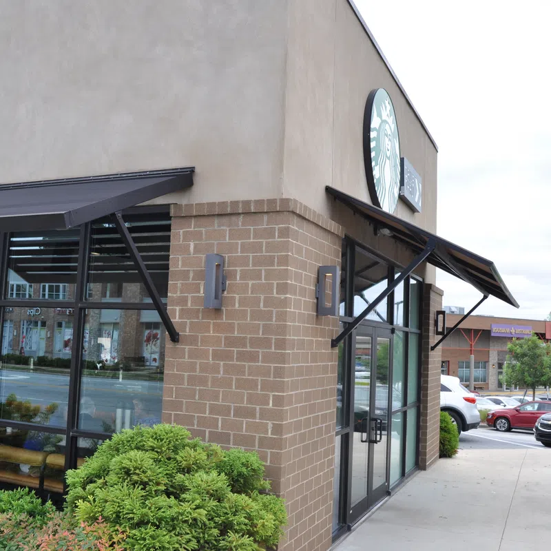 Exterior view of a Starbucks coffee shop with brick walls, large windows, awnings, and surrounding greenery on a cloudy day.
