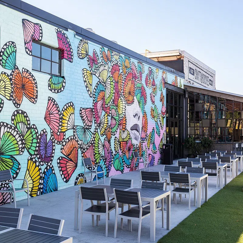 Outdoor patio with modern tables and chairs next to a colorful butterfly mural on a brick wall under clear sky