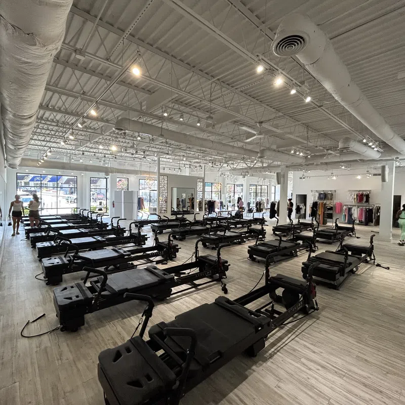 Spacious Pilates studio with multiple reformer machines, light wood floors, and people preparing for class.