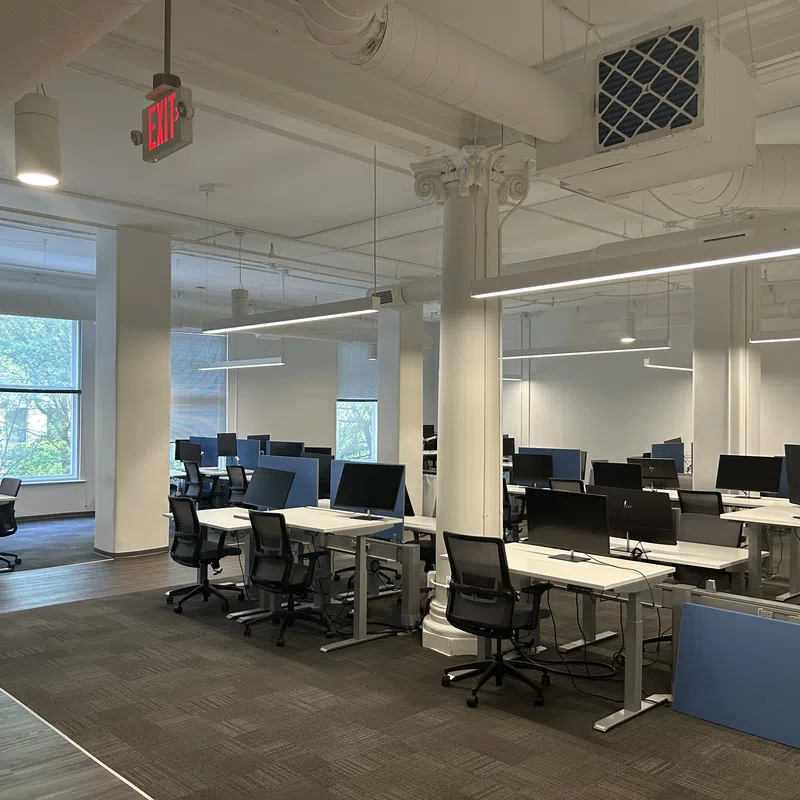 Modern office space with multiple desks, black chairs, and computer monitors under bright lighting and large windows.