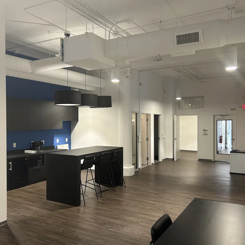 Modern office kitchen area with black island, bar stools, blue accent wall, and open hallway with wood floors.