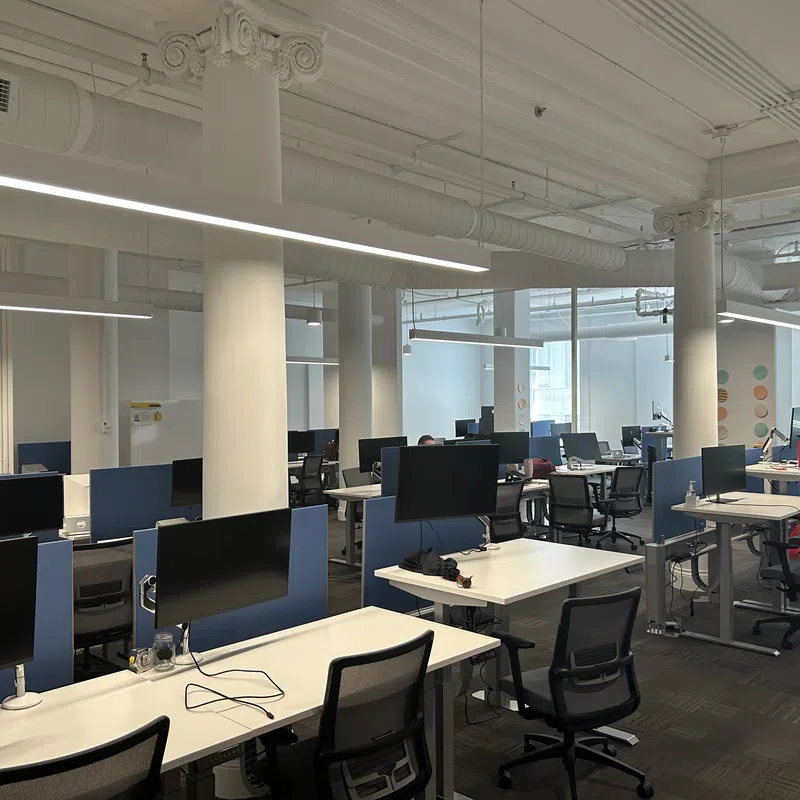Modern open office with rows of desks, computer monitors, office chairs, and white columns under bright lighting.