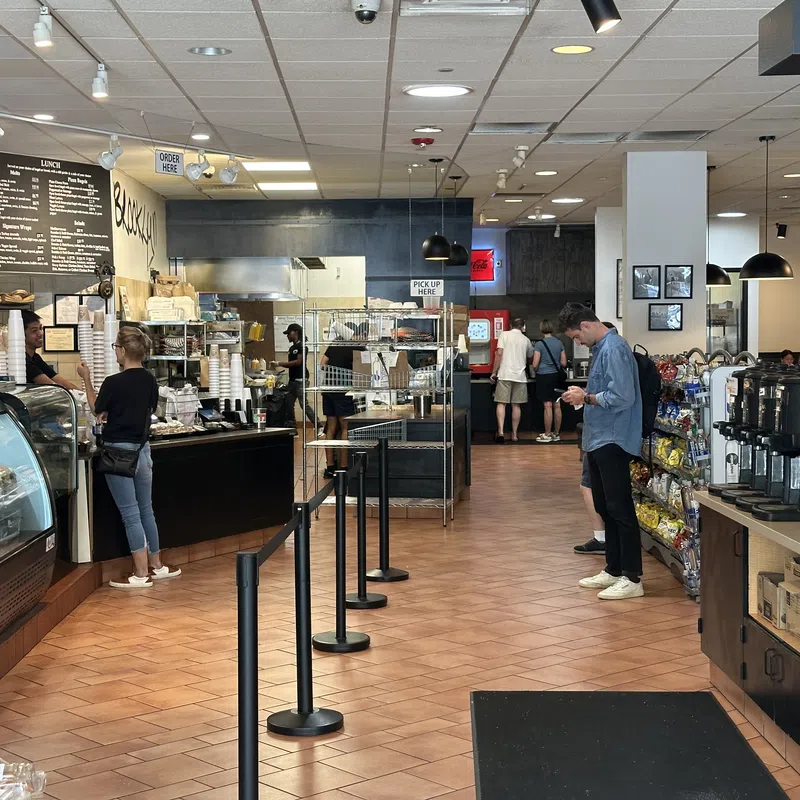 Spacious cafe interior with customers ordering, menu boards, snack shelves, and coffee station with tiled floor.