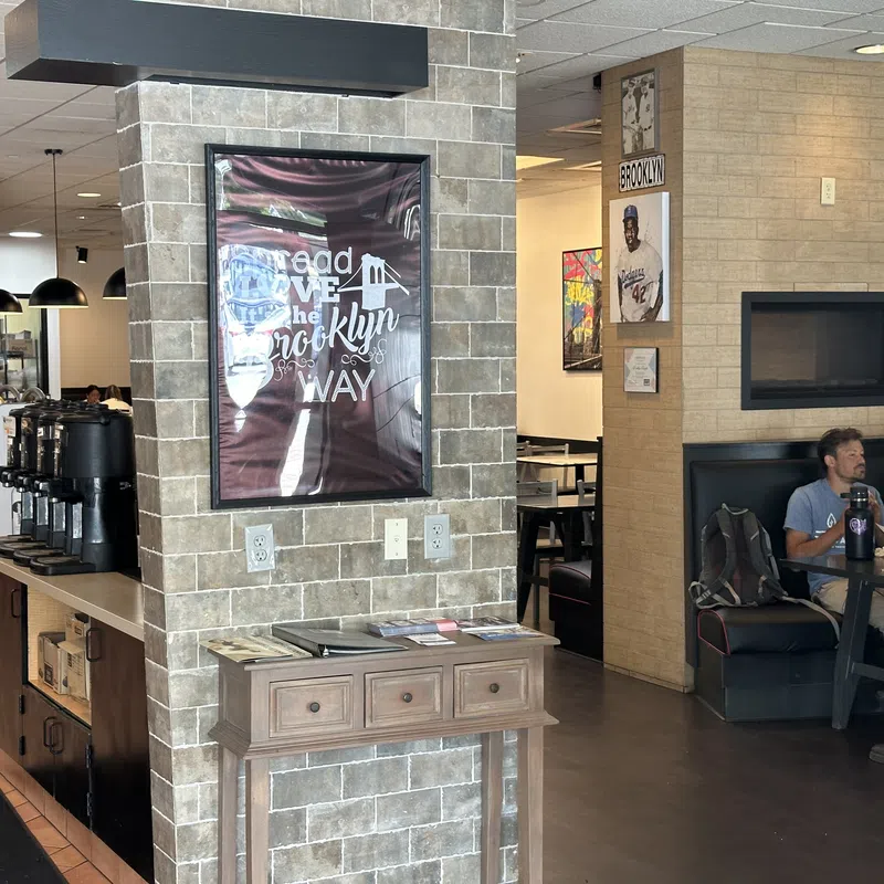 Interior of a cafe with brick walls, framed Brooklyn poster, coffee station, and a man dining at a booth.