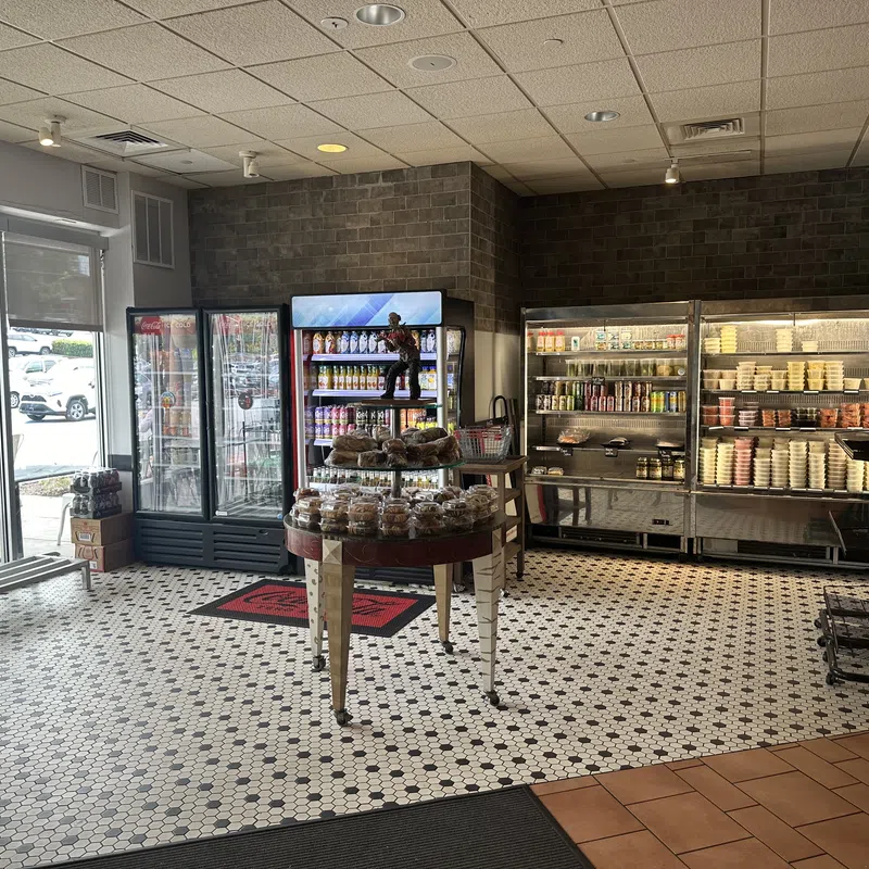 Interior of a deli with refrigerated display cases, shelves of food products, and a central table with baked goods.