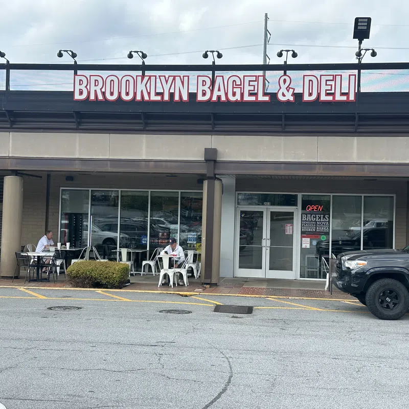 Brooklyn Bagel & Deli storefront with outdoor seating and customers under cloudy sky in a shopping plaza.