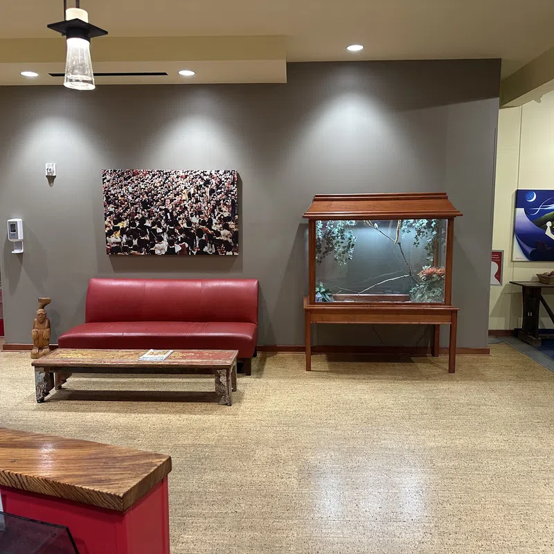 Modern waiting area with red leather couch, wooden table, glass display case, and artwork on neutral walls.