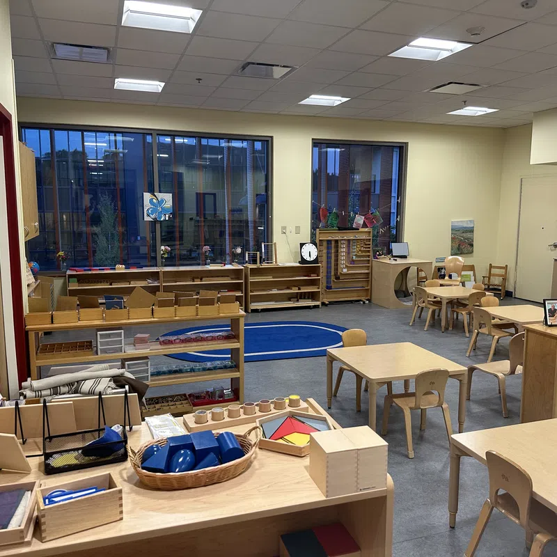 Montessori classroom with wooden tables, chairs, educational materials, and large windows letting in natural light
