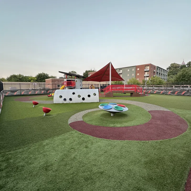 Empty modern playground with artificial turf, red and gray play elements, and a shaded canopy near buildings.