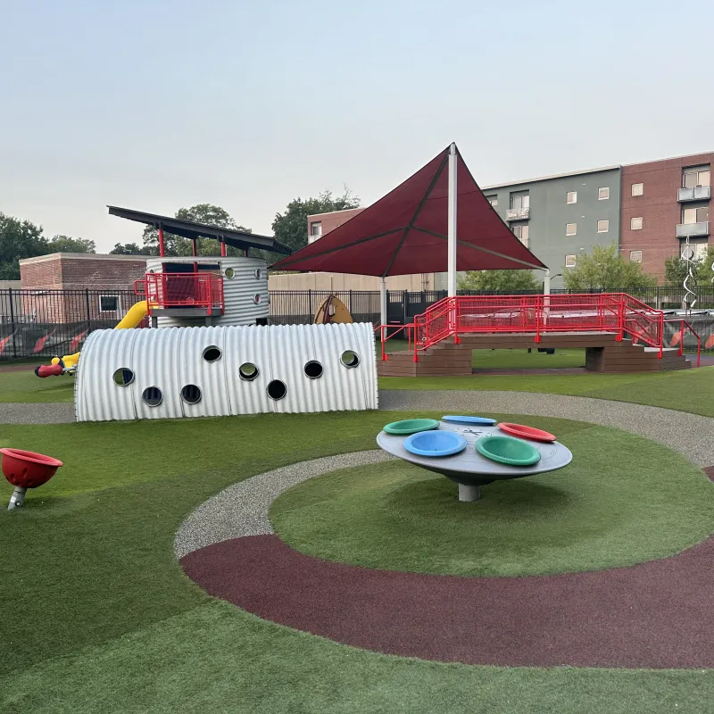Modern outdoor playground with colorful equipment, climbing tunnel, shaded area, and rubber safety flooring under clear sky
