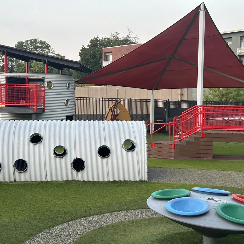 Modern playground with a large red shade canopy, corrugated tunnel, colorful spinning play equipment, and a slide.