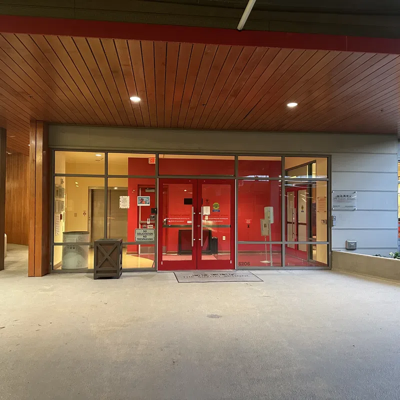 Entrance to a modern building with red doors, glass panels, wooden ceiling, and concrete floor at dusk